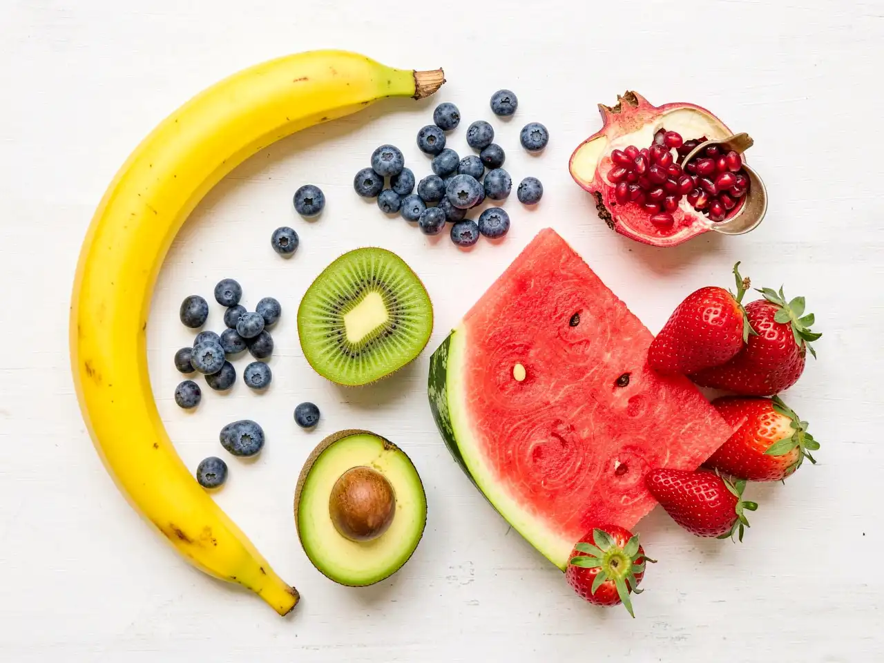 A colorful flat lay of fresh, whole fruits including a banana, blueberries, sliced kiwi, watermelon wedge, strawberries, and half an avocado on a light background