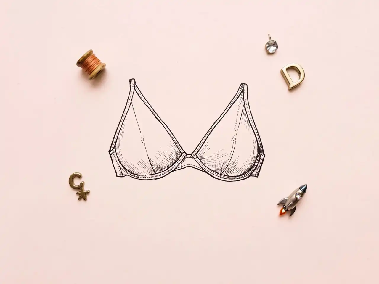 A vintage-style line drawing of a bra on a light pink background, surrounded by a spool of thread, letter charms, a rhinestone, and a tiny rocket