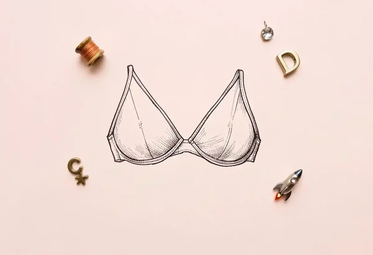A vintage-style line drawing of a bra on a light pink background, surrounded by a spool of thread, letter charms, a rhinestone, and a tiny rocket