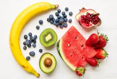 A colorful flat lay of fresh, whole fruits including a banana, blueberries, sliced kiwi, watermelon wedge, strawberries, and half an avocado on a light background