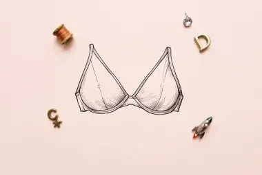 A vintage-style line drawing of a bra on a light pink background, surrounded by a spool of thread, letter charms, a rhinestone, and a tiny rocket