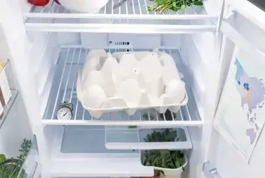 An open refrigerator with a white egg carton prominently on the shelf, symbolizing the American practice of refrigerating eggs for food safety.