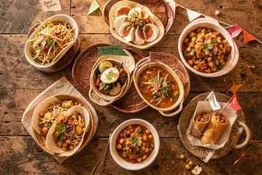 Overhead view of various cultural potluck dishes with small flags and labels on a rustic table